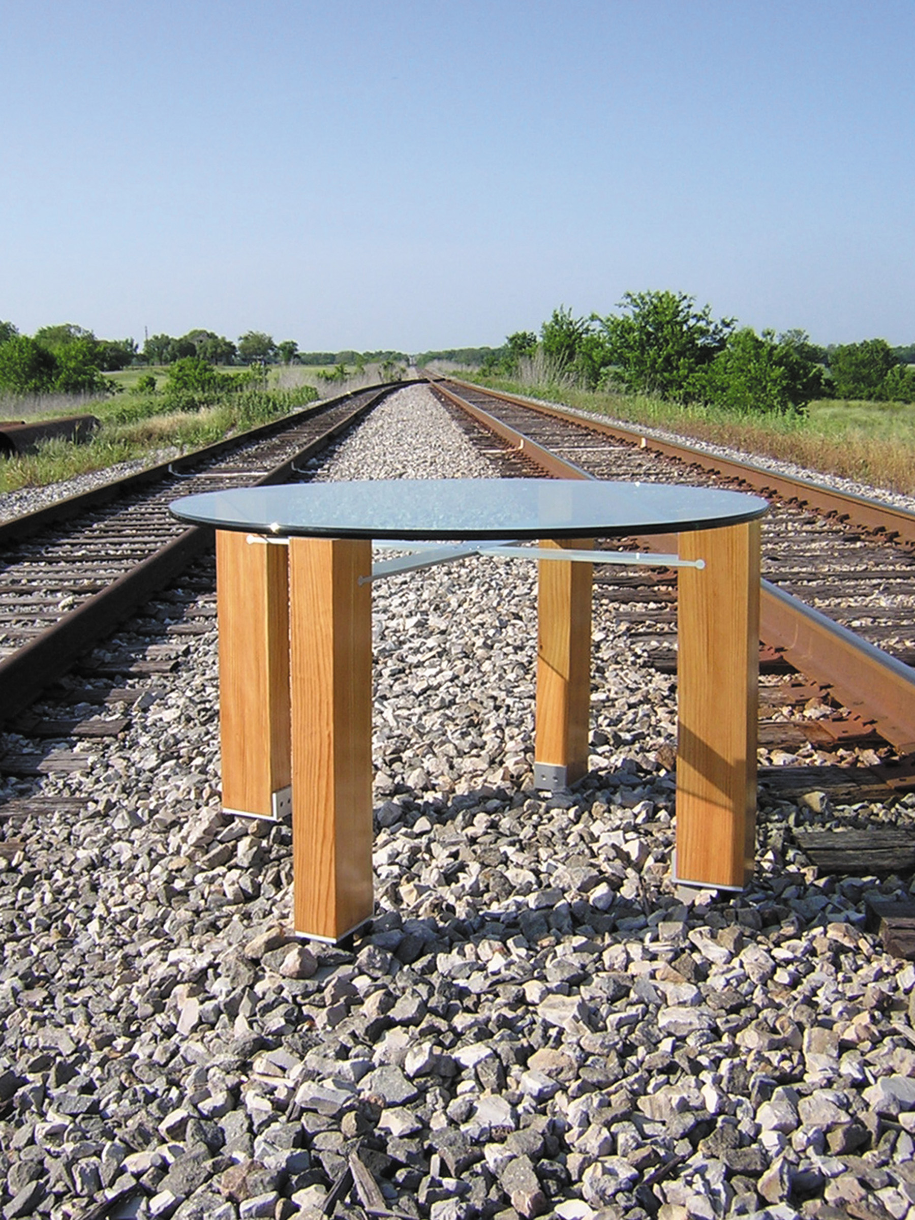 Contemporary table with round glass top, antique longleaf pine legs, and aluminum stretchers that connect the legs. The aluminum fits into the longleaf pine legs with provocative joinery. The longleaf pine has a natural finish. The table is photographed on the railroad tracks outside our studio in Texas.